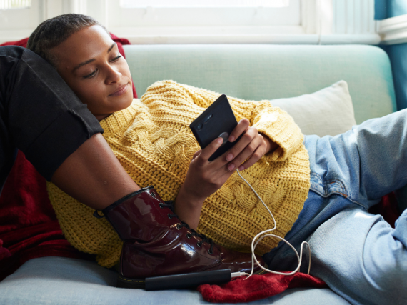 A Black woman relaxes against a friend's legs on the sofa while scrolling through her phone. 