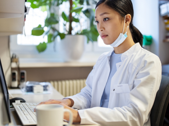 A pharmacist sits alone at a computer with their mask sitting below their face as they work on prescriptions. 