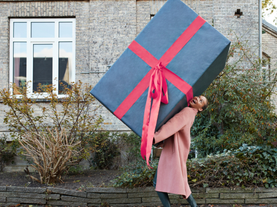 A woman walks down the street holding a comically large box. 