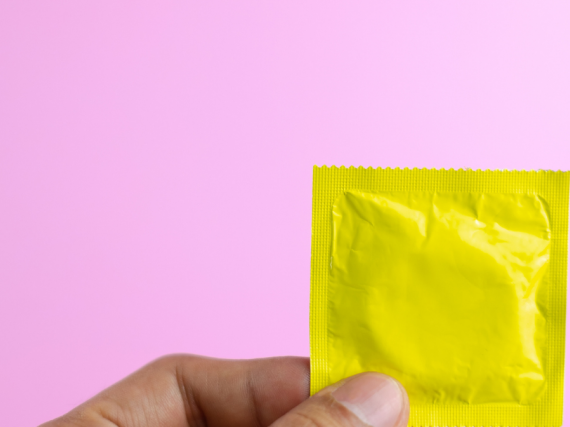 A hand holds a condom in a bright yellow wrapper against a light pink background. 