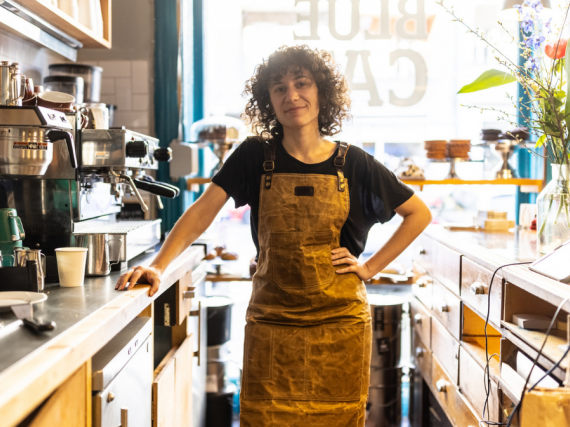 A person stands confidently behind the counter of the coffee that they work in. 