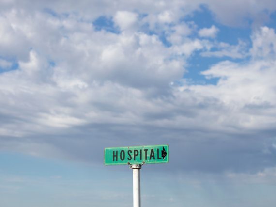 A green sign with the word hospital sits in the middle of a field against a blue sky. 