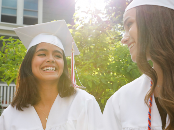 Two girls wearing white high school graduation gowns and smile. 