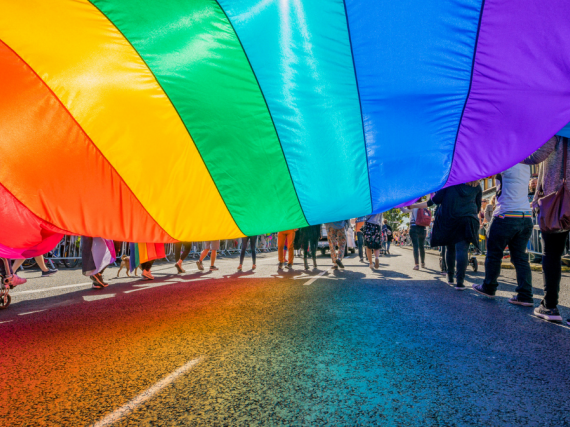 A photo from underneath a massive pride flag being carried by many people during a parade. 