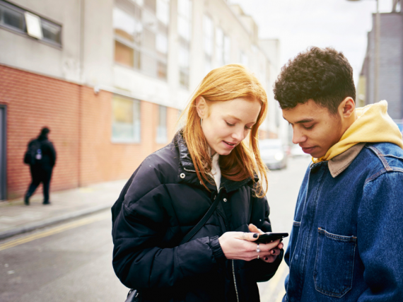 A young teen couple stands in the street and looks at a cell phone. 