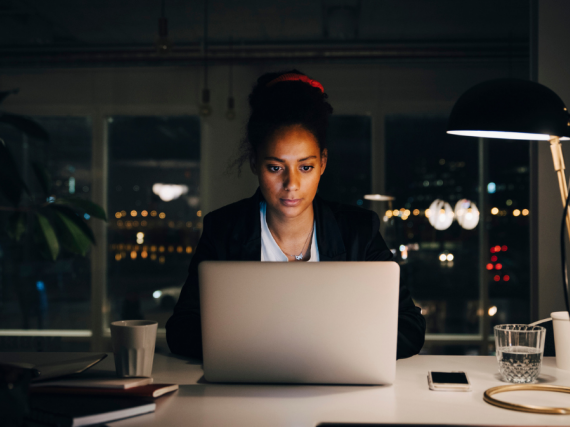 A woman sits at a desk on her computer late at night with only one light to work by. 