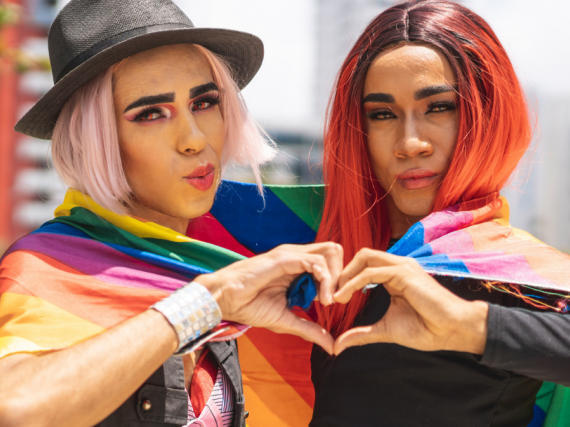 Two queer people wrapped in a rainbow flag make a heart with their hands. 