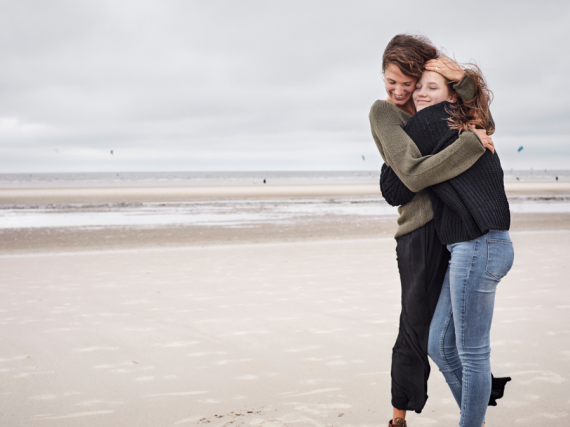 A mother and daughter laugh and hug while standing on a beach. 