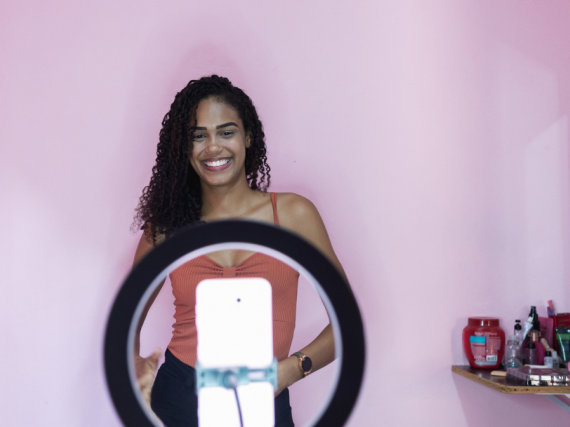 A woman poses in front of a pink wall with a phone set into a ring light in front of her, filming. 