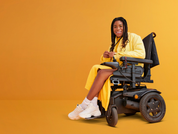 Against a yellow background, a Black woman dressed in yellow and sitting in a wheelchair smiles broadly.