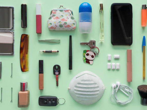 A green table with the contents of someone's bag laid out. Items include a mask, phone, comb, bobby pins, headphones, and more. 