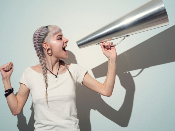 A woman holds a silver megaphone up to yell into against a blue background. 