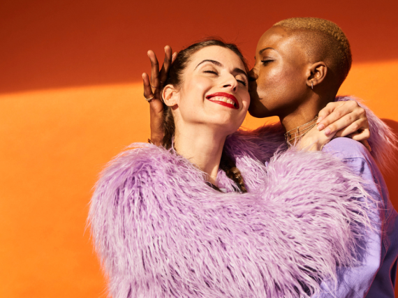 A woman kisses another woman's face while they hug against a bright orange background.