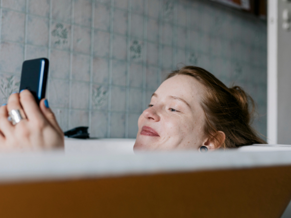 A woman relaxes in a bath with a smile as she looks at her phone. 