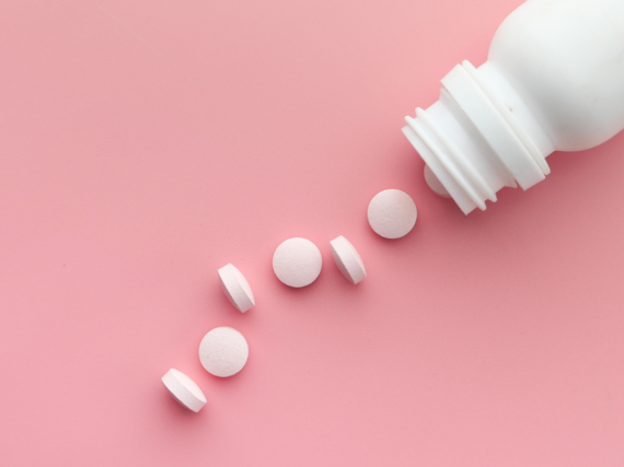 A blank white bottle with small white pills spilling out on a pink background. 