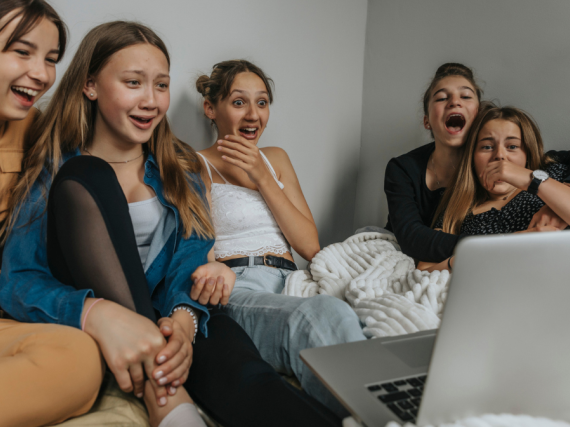 A group of 5 teen girls watch TV together and laugh wildly. 