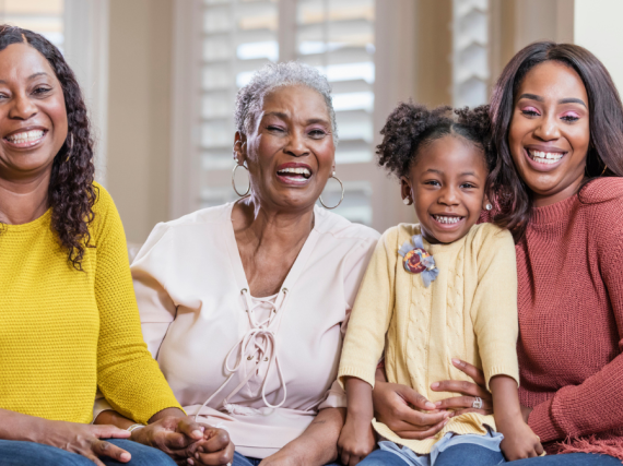 Four generations of Black women sit on a sofa and smile at the camera.