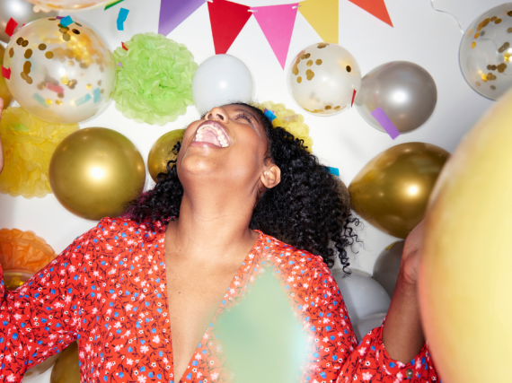 A woman stands and throws her head back laughing while surrounded by balloons. 