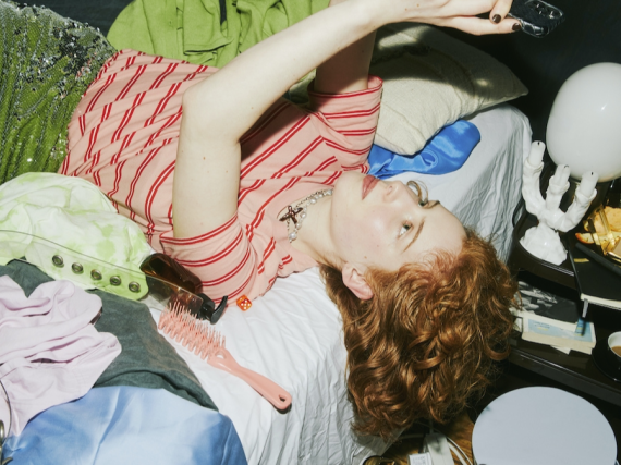 A young woman scrolls on her phone while laying on a messy bed. 