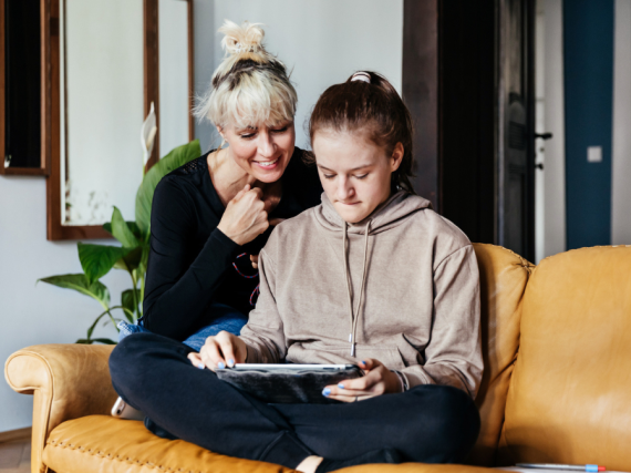 A mother and her teenage daughter sit on the sofa with an ipad. 
