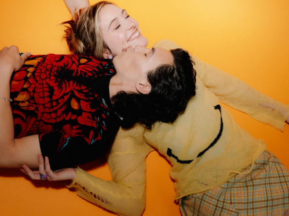 A teen couple lays on the ground and laughs. The man kisses the woman's cheek as his head rests on her chest. 