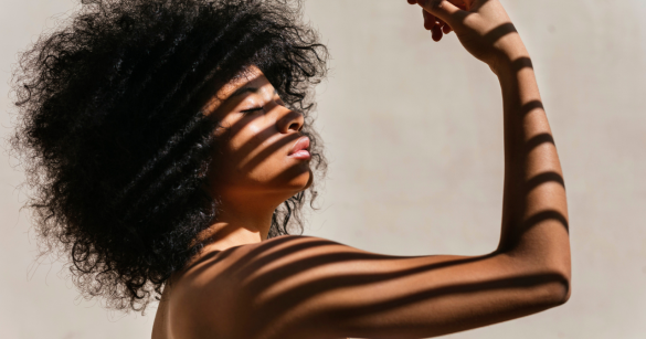 A Black woman curls her bicep while the sun shines through blinds making shadows across her body. 